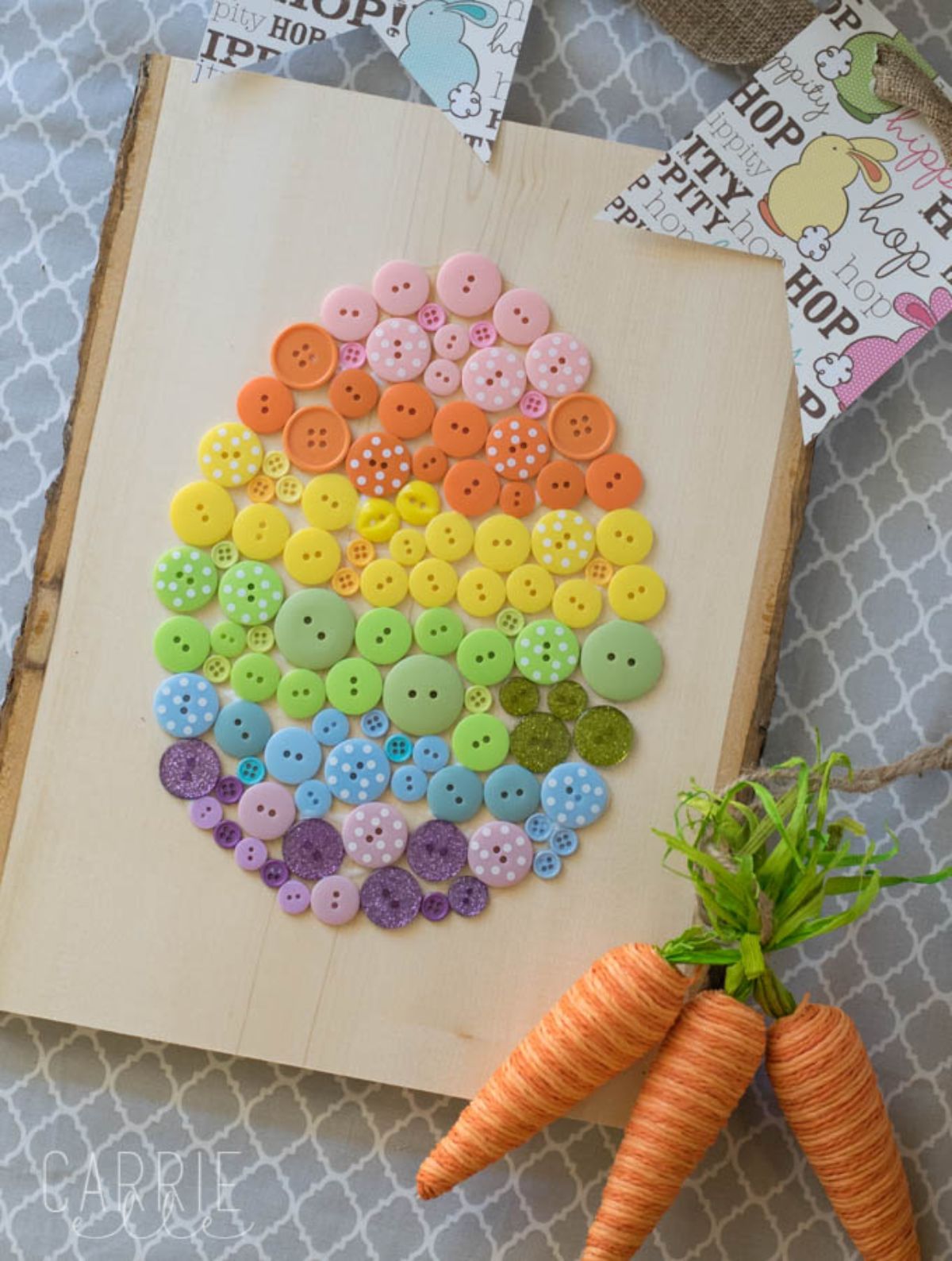 A striped egg made of different colored buttons is stuck to a canvas sheet. Next to it sits 3 material carrots