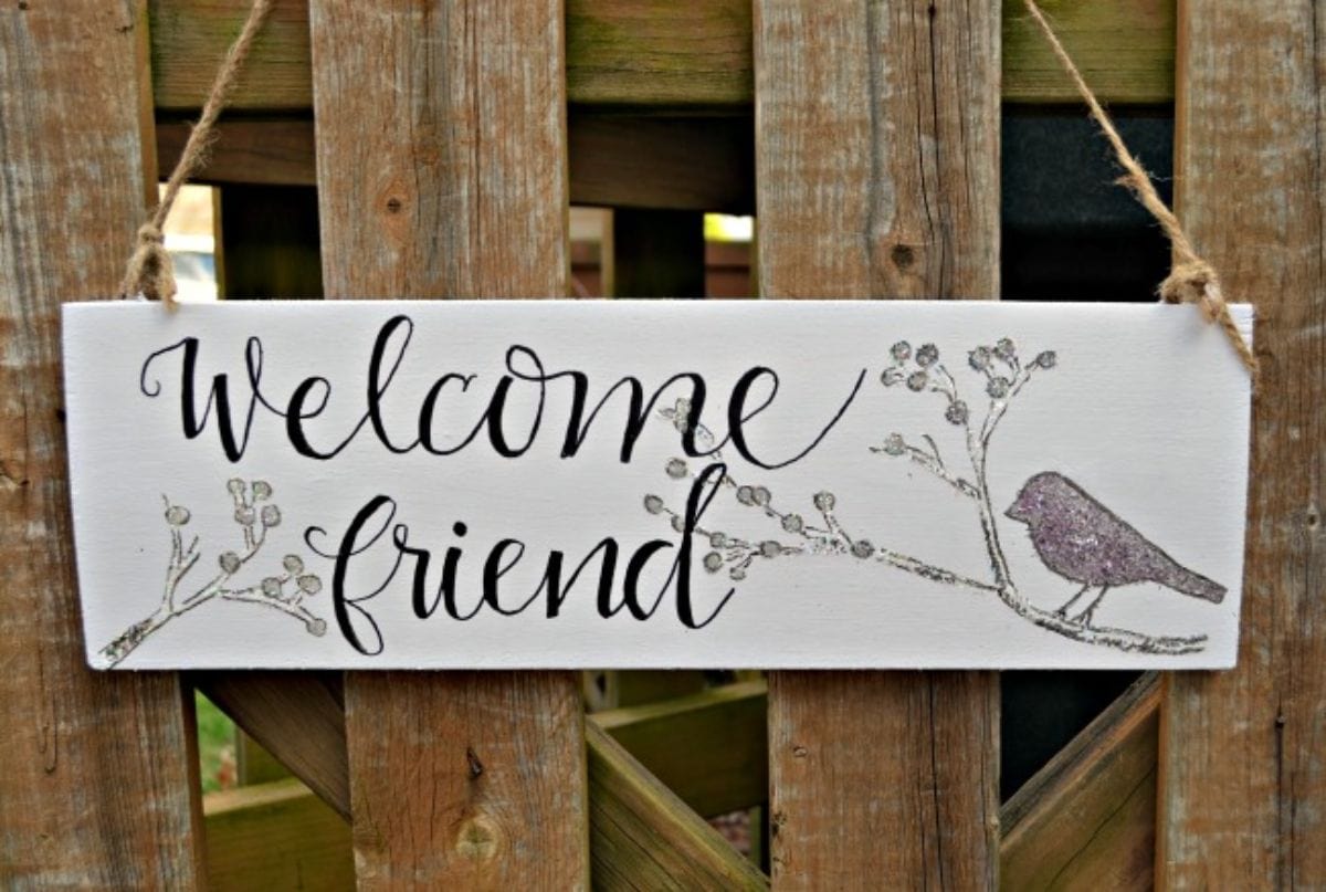 A painted sign is hanging from a crate. Words say "Welcome friend" and there is a drawing of a bird to the right