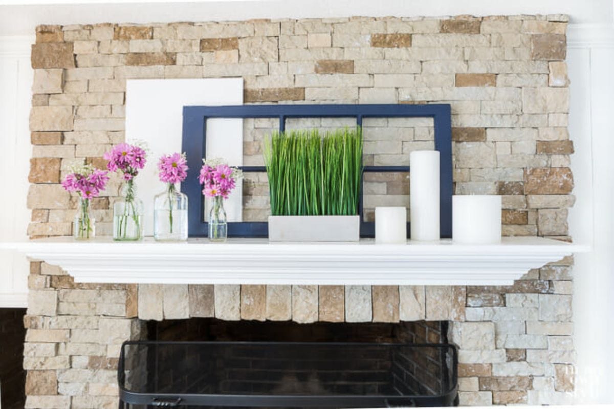 A mantelpiece is on a brick wall. There are 2 empty picture frames, some flowers in vases and a white ceramic pot filled with green stems