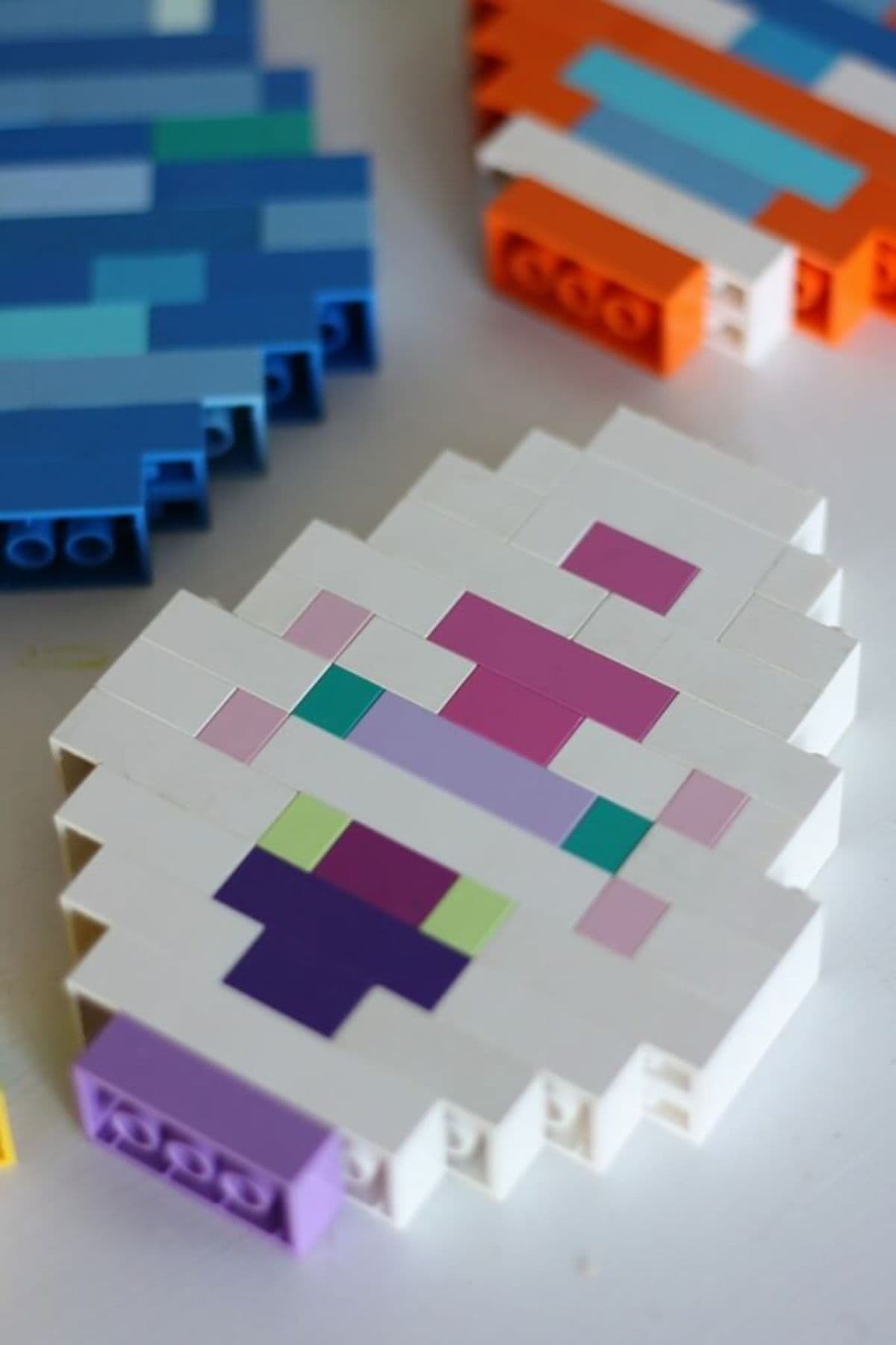 A blue egg, purple and white egg, and orange white and blue egg all made of lego are laying down on the surface