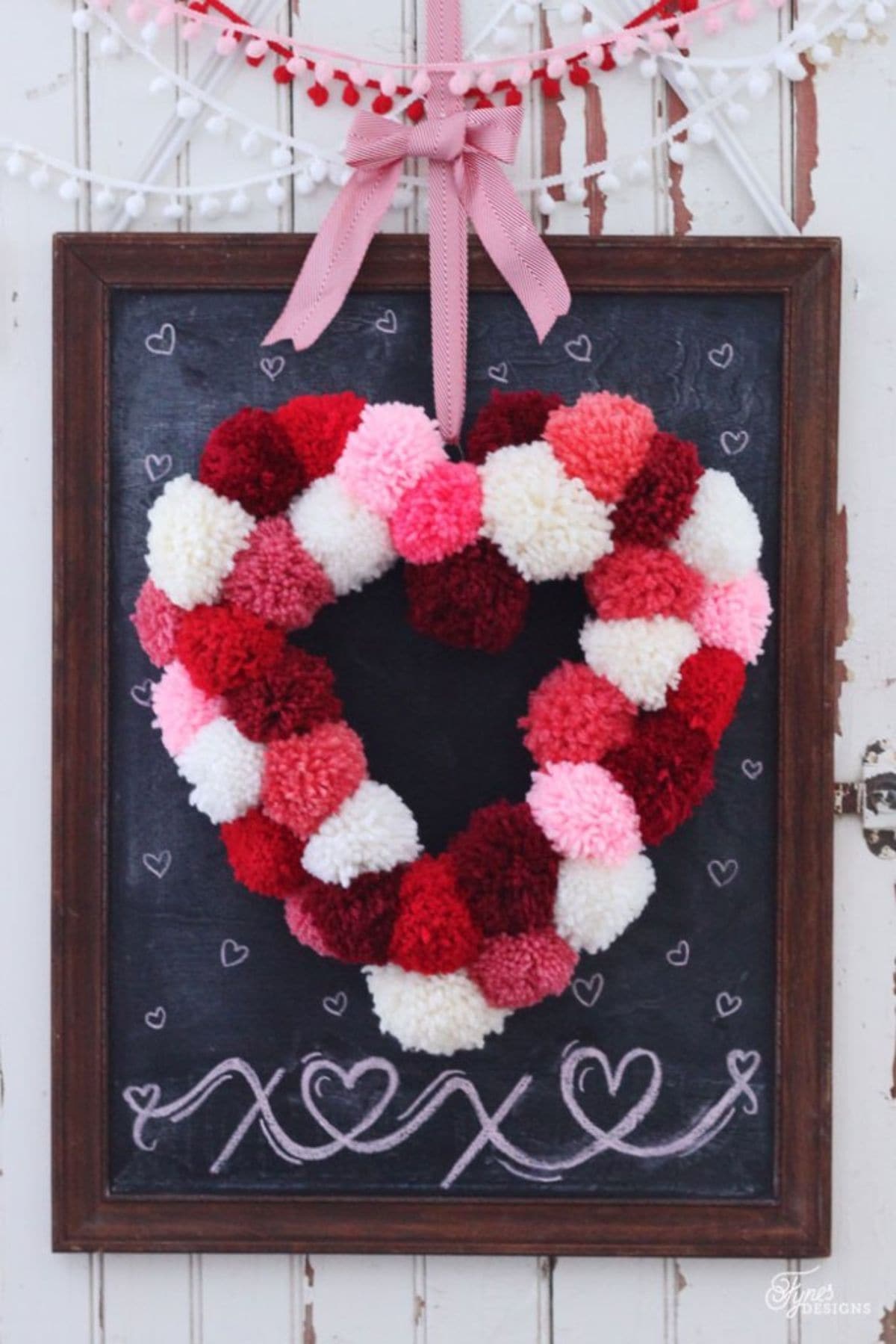 Hanging from a framed blackboard by a pink ribbon, is a heart made out of pom poms in reds, whites and pinks.