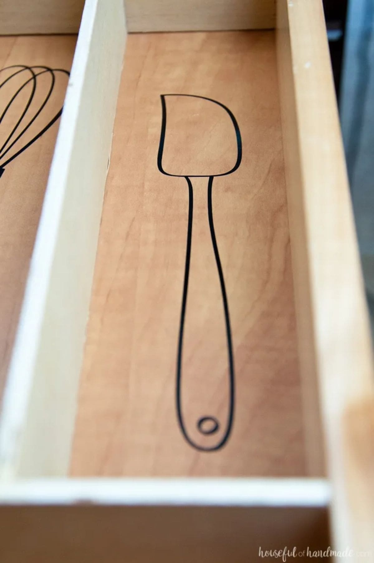 A close up of a section of a drawer with wooden dividers. There is a black pen drawing of a spatula on the bottom of the drawer