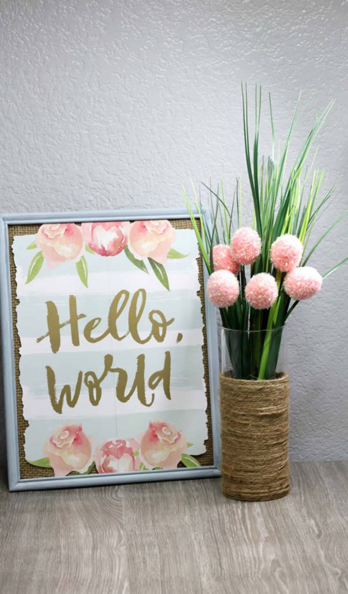 Next to a painted sign saying "hello world!" is a thin glass vase filled with tulips. Twine is wrapped around the bottom half of the vase