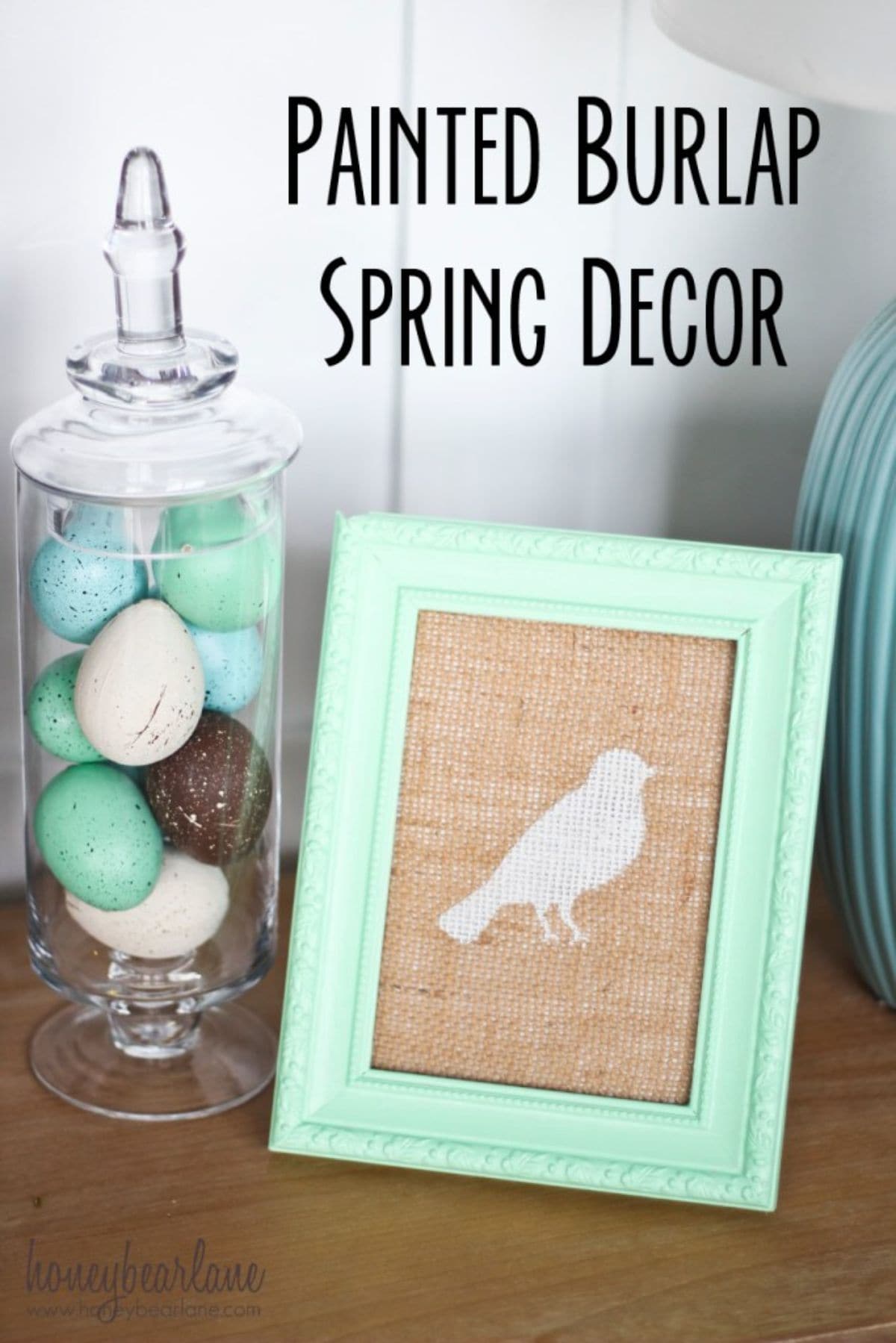 On the left of the picture is a glass jar full of colored eggs. On the right is a burlap sheet with a green painting of a bird on it, it is framed in green