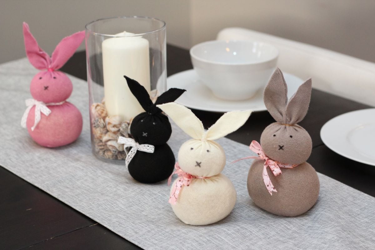 4 bunnies, pink, black, cream and grey sit on a table runner in front of a candle in a glass lantern