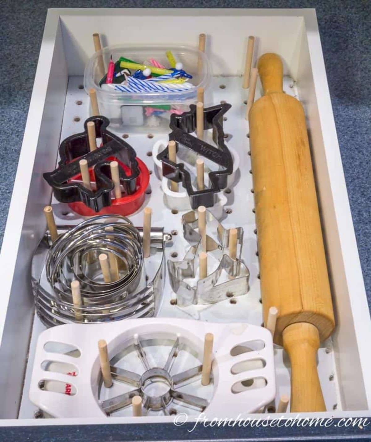 The inside of a white kitchen drawer is shown. The bottom has been lined with a peg board adn wooden dowels are sticking up at random intervals. Cookie cutters and other backing suppliesa re fitted around the dowesl. A rolling pin is on the right hand side