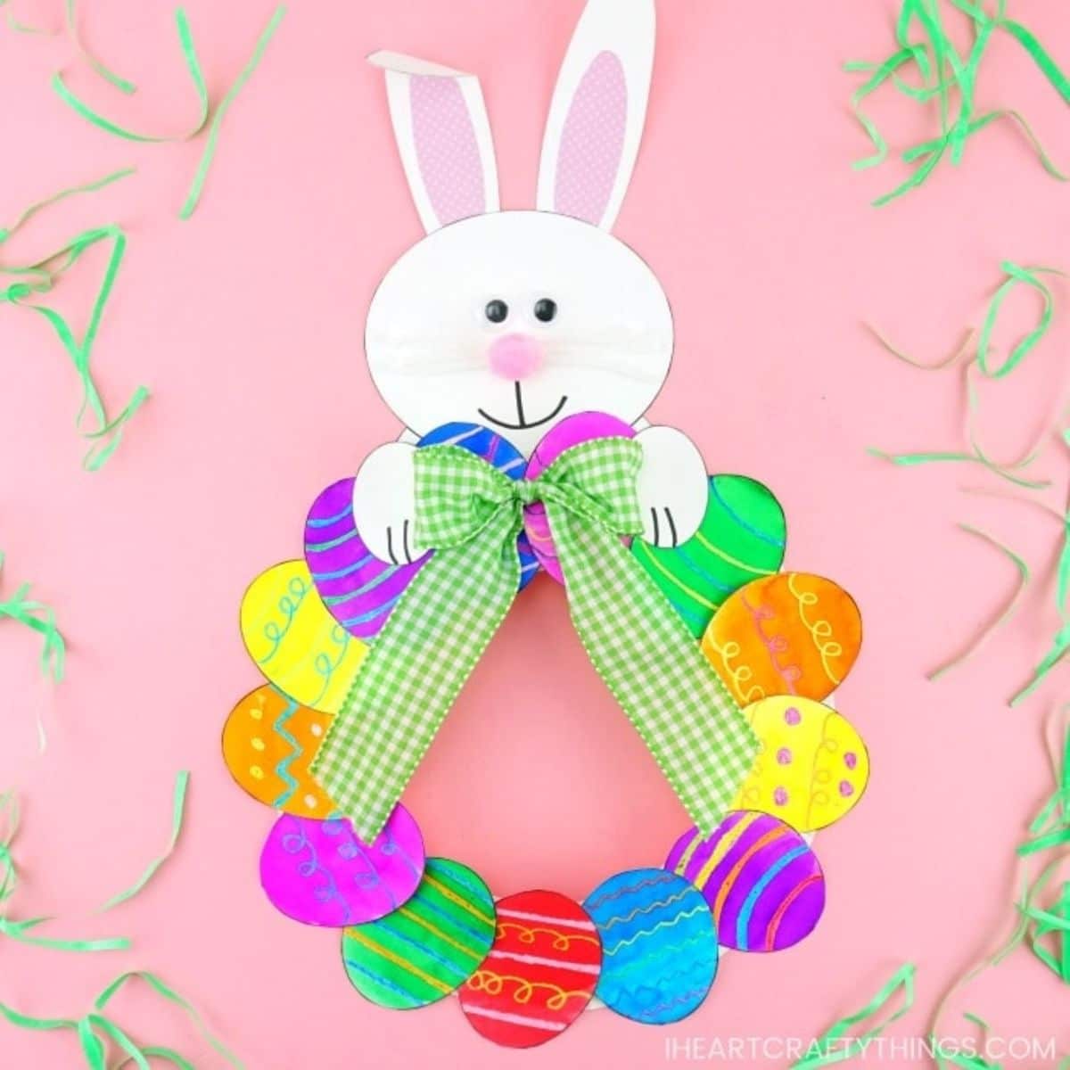 On a pink background a curcle made of colored paper eggs sits underneath a bunny's head