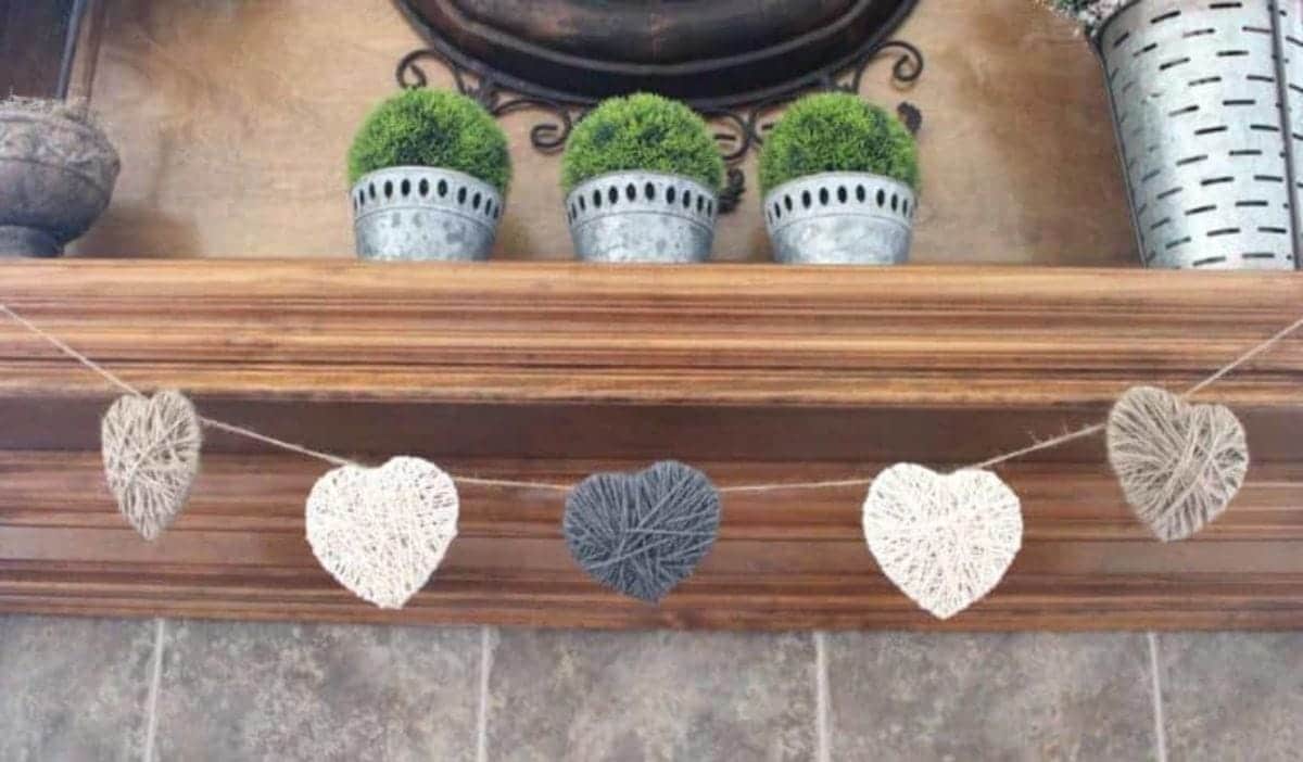 A wooden mantelpiece is decorated with 3 metal plant pots filled with gree, bushy plants. Hanging from the mantel is a garland of yarn hearts in greys and whites