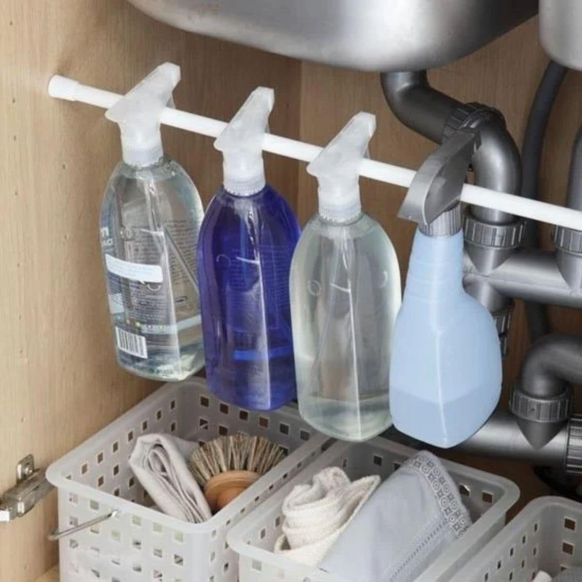 under the kitchen sink a tension rod has been fixed just under a stainless steel bowl. Hanging on teh rod are cleaning product bottles. Underneath are white baskets for other cleaning supplies