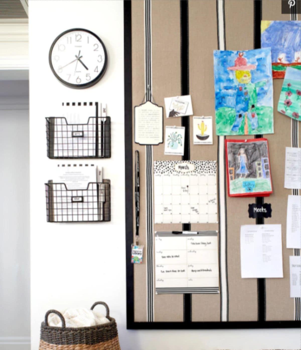 A kitchen wall is shown, with a clock at the top, and two shallow baskets underneat, holding paperwork. On the righ hand side is a display board with artwork and calendars on it.