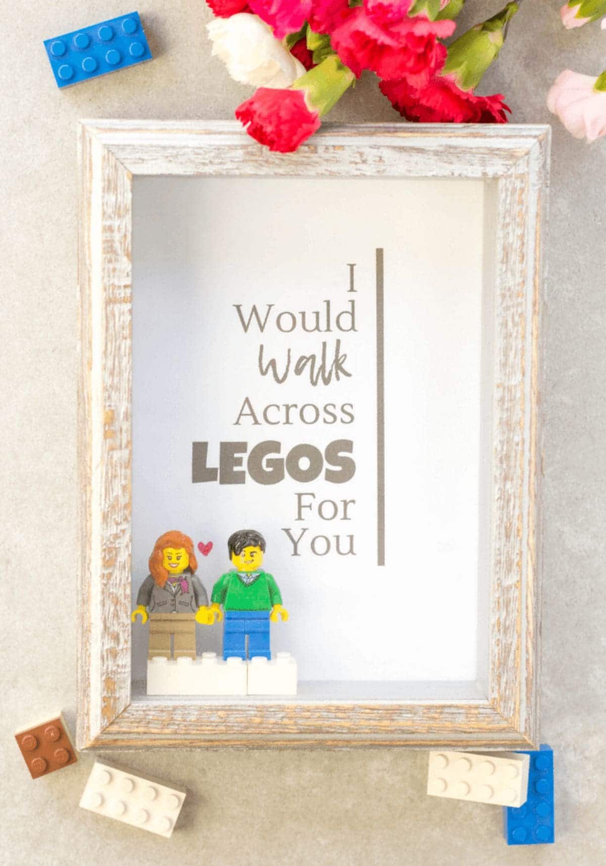Against a cream background wit hlego pieces scattered around it is a whit wooden box frame. Inside are a male and female lego figure. Above them is the text "I would walk across legos for you"