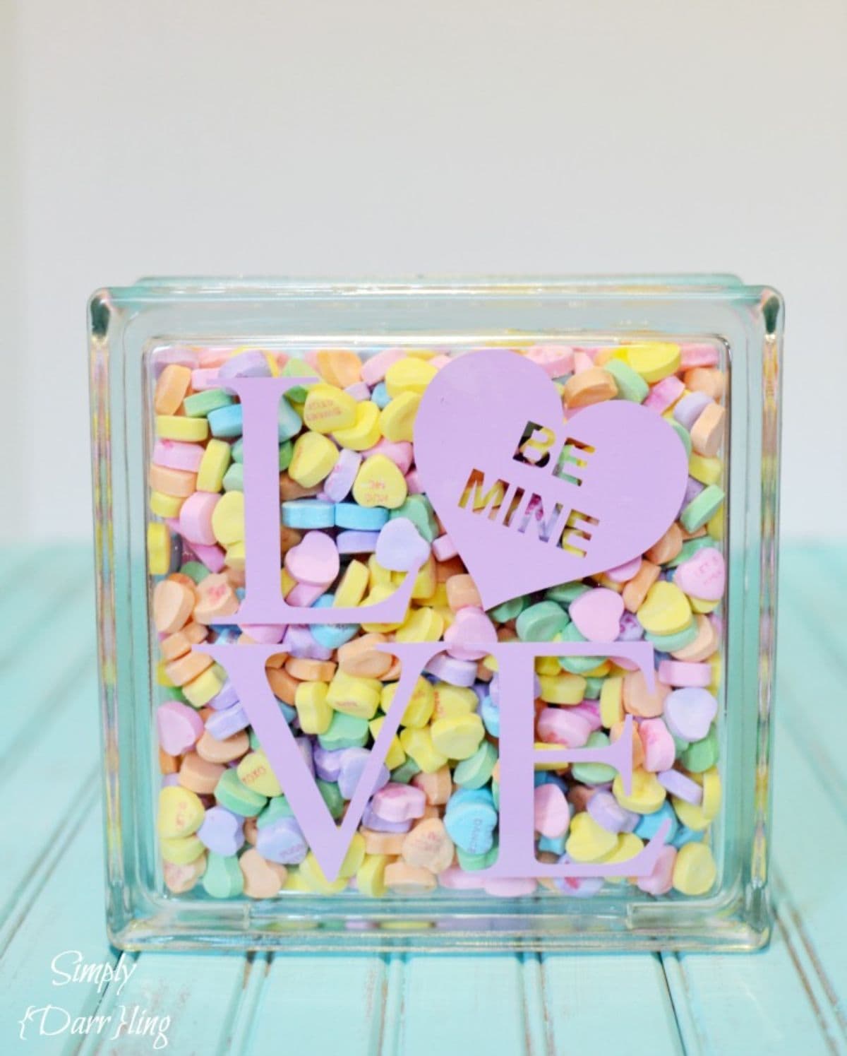 on a turqoise blurred table is a perspex block with teh words "LOVE" on the front in pink. The O is a heart. The block is full of multi-colored candy hearts