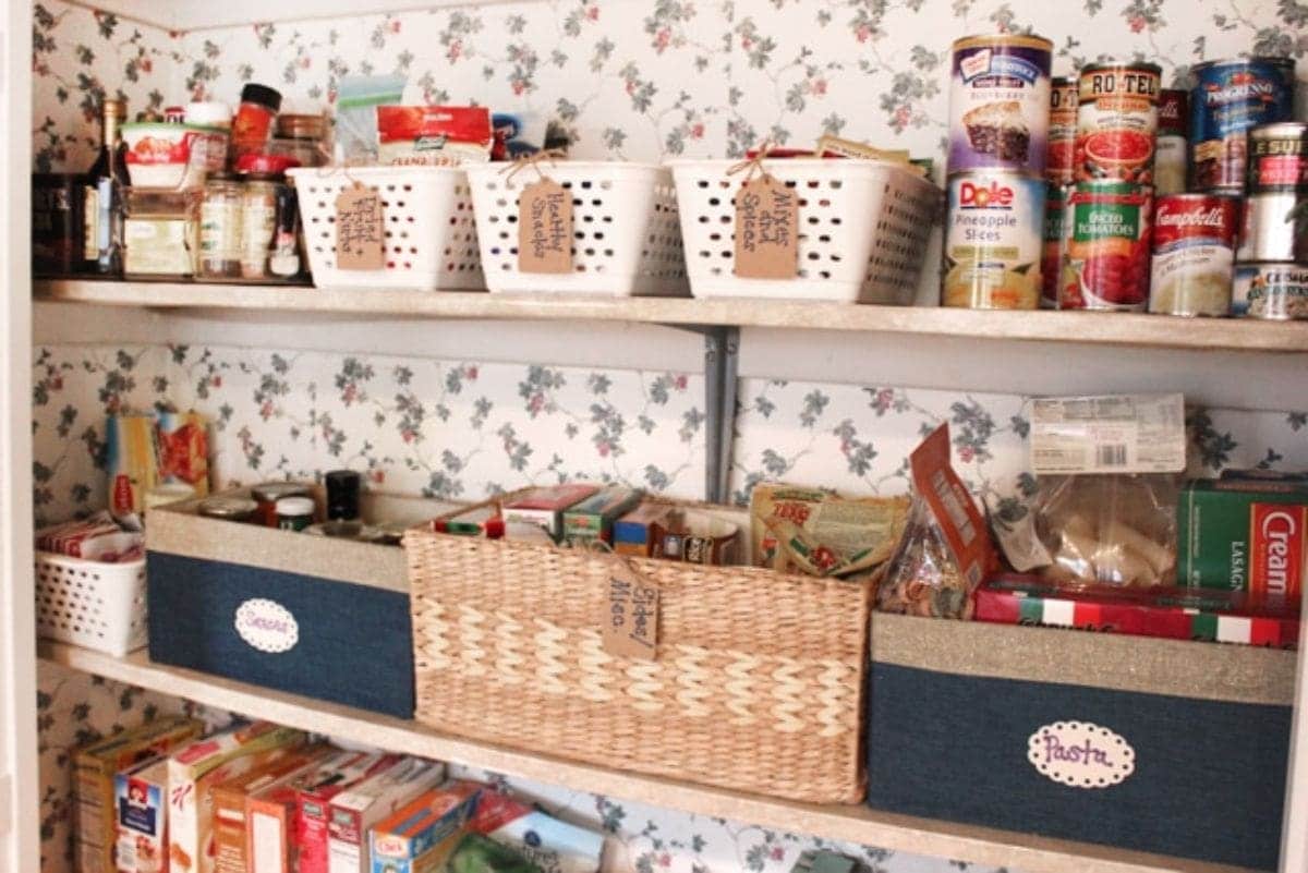 flowery wallpaper with 2 wodden shelves. On each shelf are different baskets with parcel labels attached to them. They are filled with pantry goods