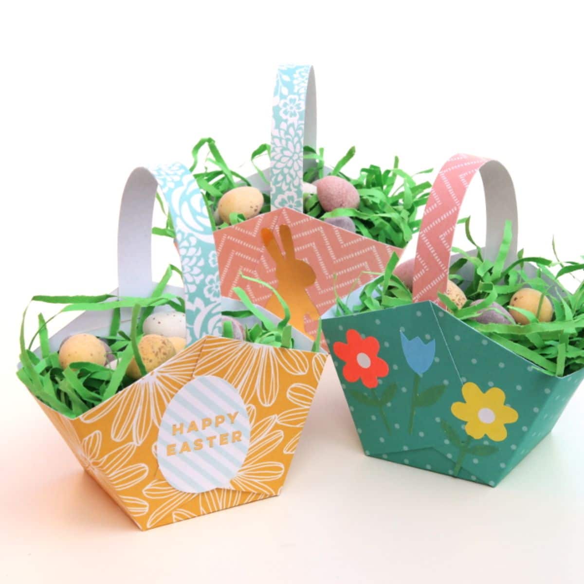 3 paper baskets colored in green, orange and pink and decorated with flowers contain fake grass and chocolate eggs