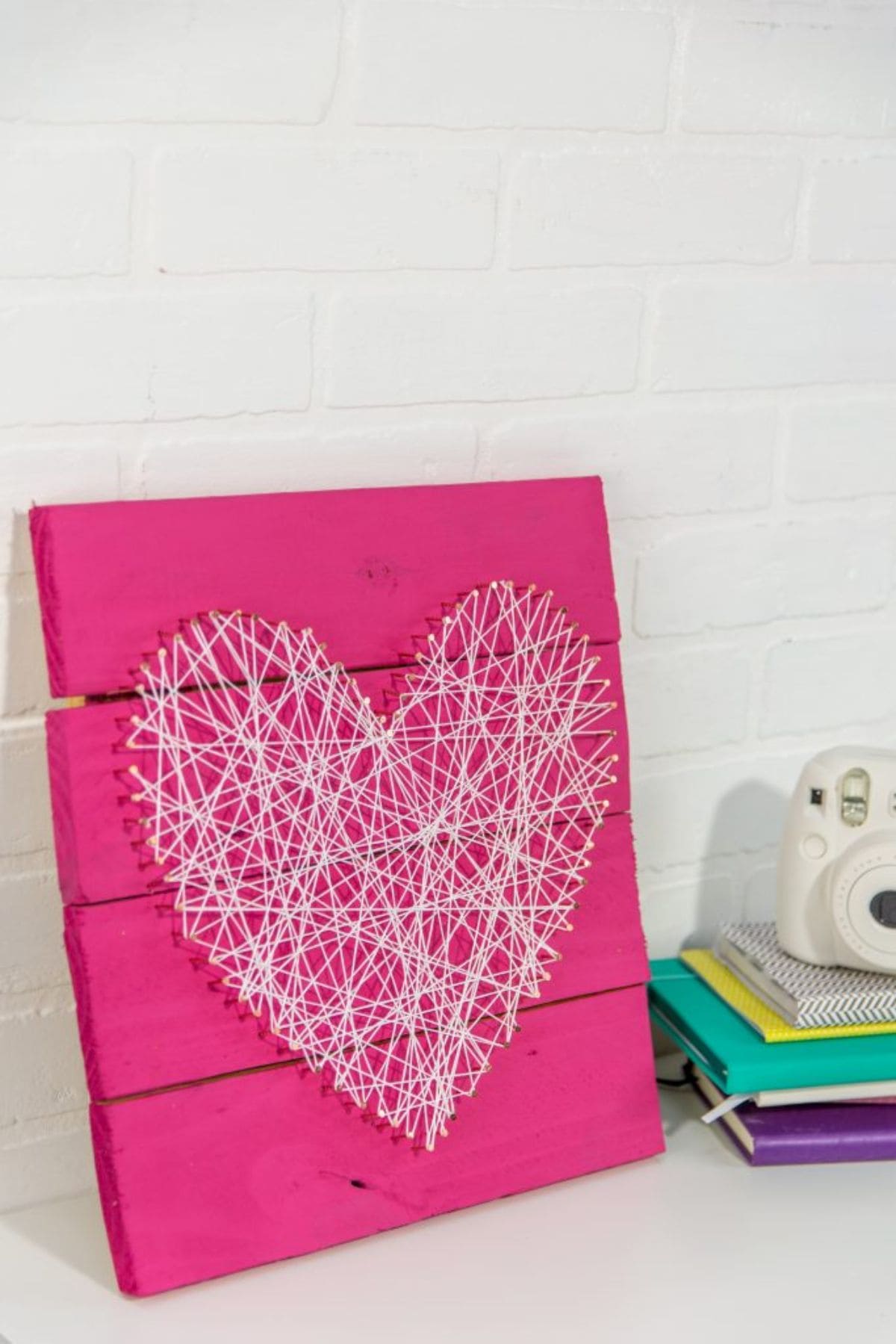 Resting up against a cream wall is a pink wooden board. On the front of it is a heart made from string and nails in white.