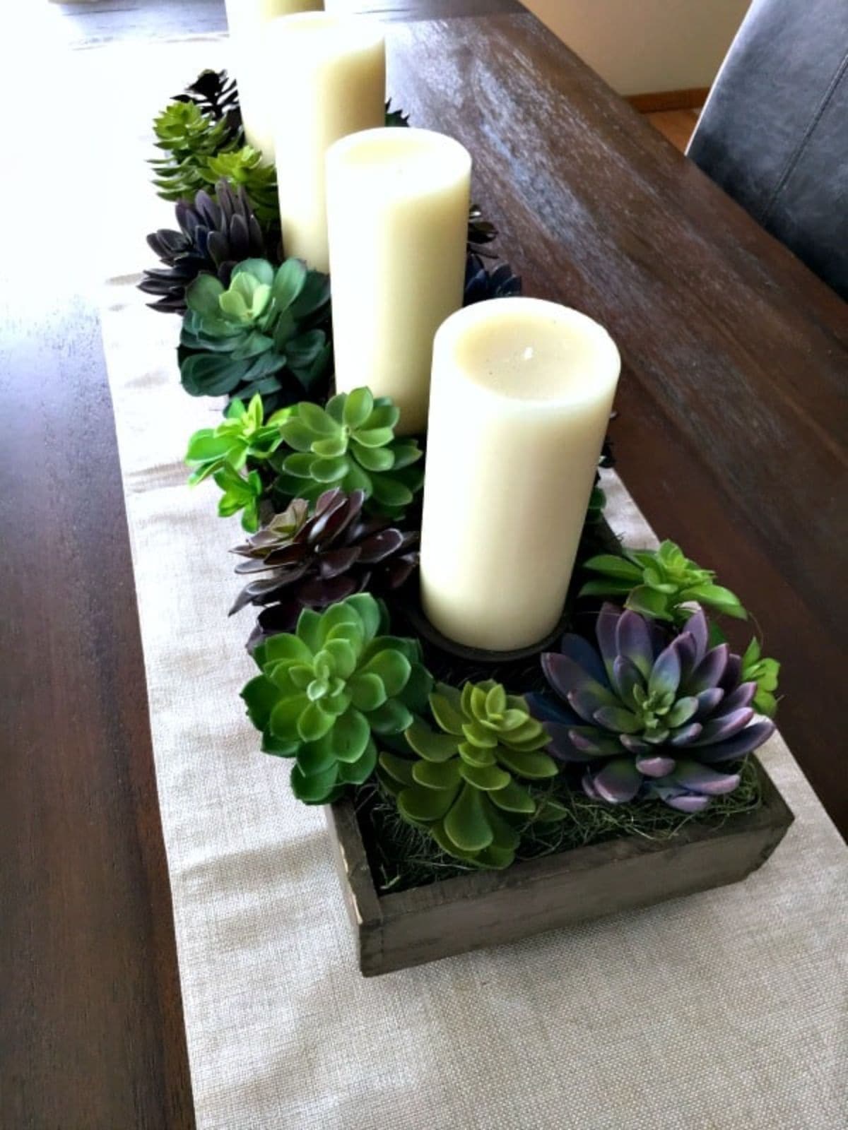 Sitting on a wooden table and a white table runner are 3 church candles surrounded with green wreaths