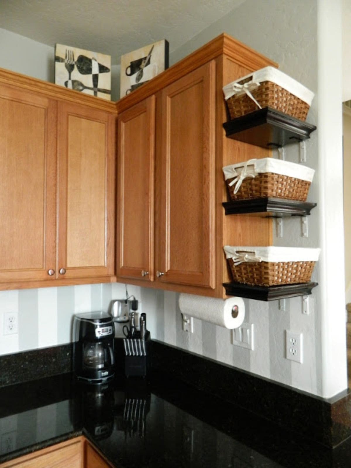 THe corner of a kitchen, showing light wood untits. On the edge of the unit 3 shelves have been fixed and wicker baskets sit on each of these shelves.