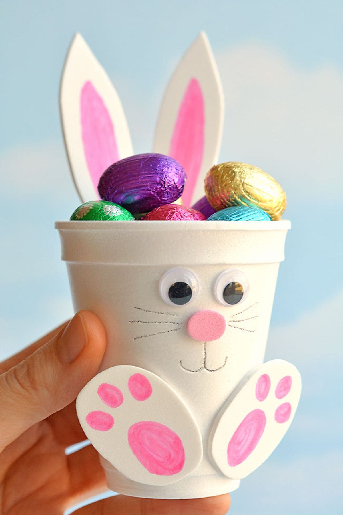 A hand holds a white foam cup decorated to look like a bunny with foil-covered chocolate eggs inside