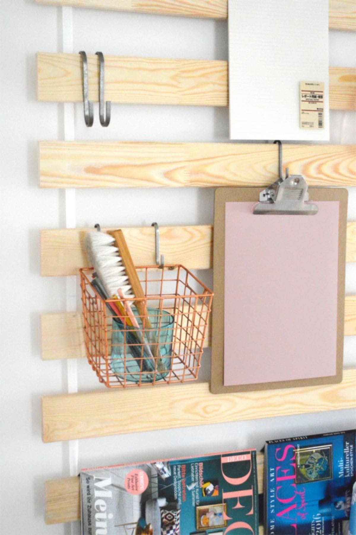 A section of a wooden slatted headboard. Hanging from the slats are hooks. Hanging fro the hooks is a basket with stationery in it and a clipboard