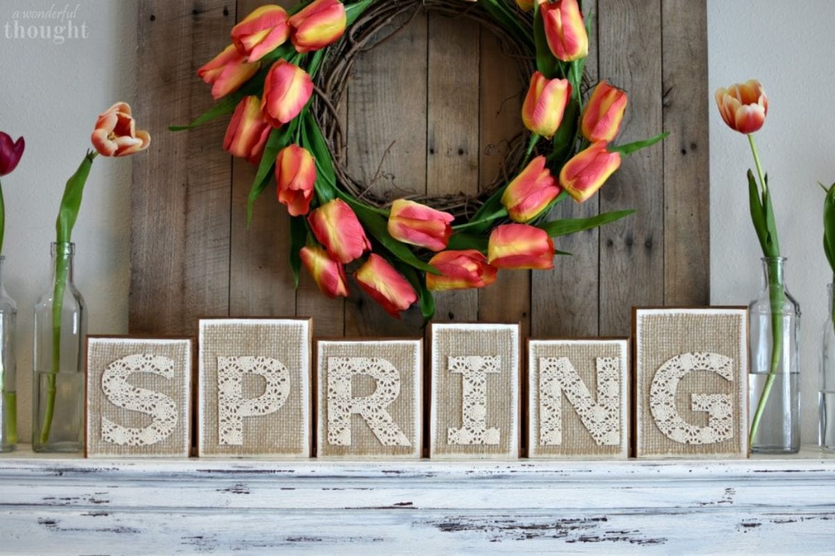 under a wreath made of orange tuips sit 6 wooden blocks on a mantelpiece spelling out "Spring"