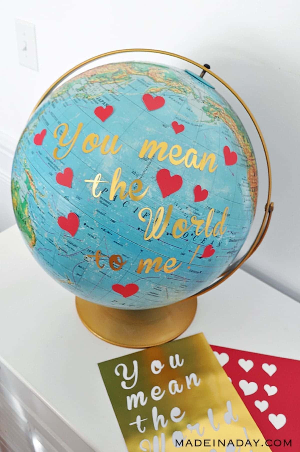 A globe with a gold stand sits on a white table. The globe is covered in red hearts and the tgold text "You mean the world to me"