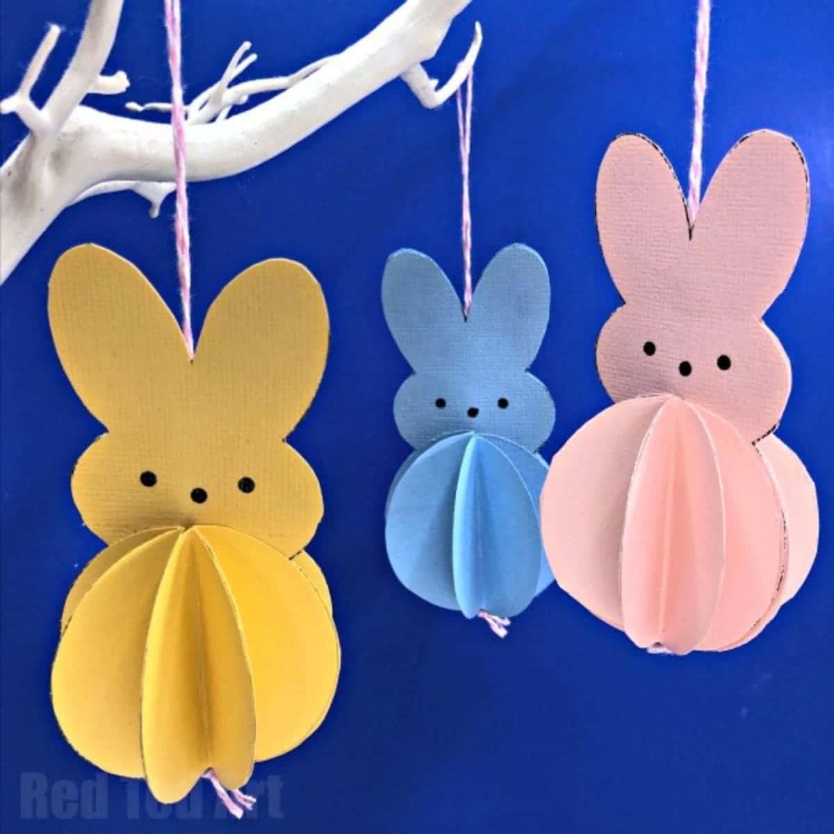 3 paper bunny shapes in orange, blue and pink hang from a white branch against a blue background