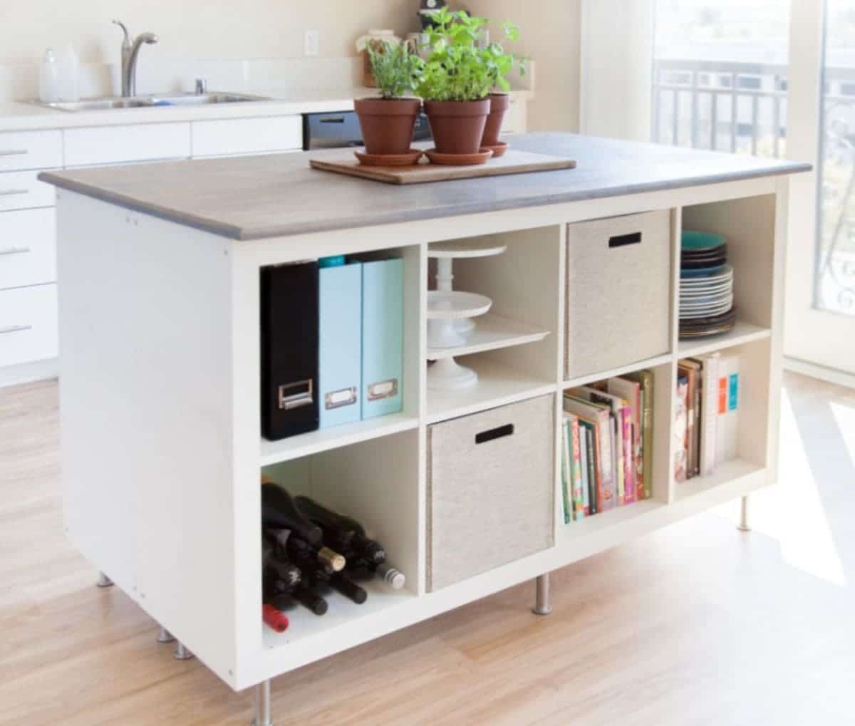 A white IKEA Kallax unit sits on coasters and is filled with wine bottles, folders, plates and baking trays. On top are some potted plants