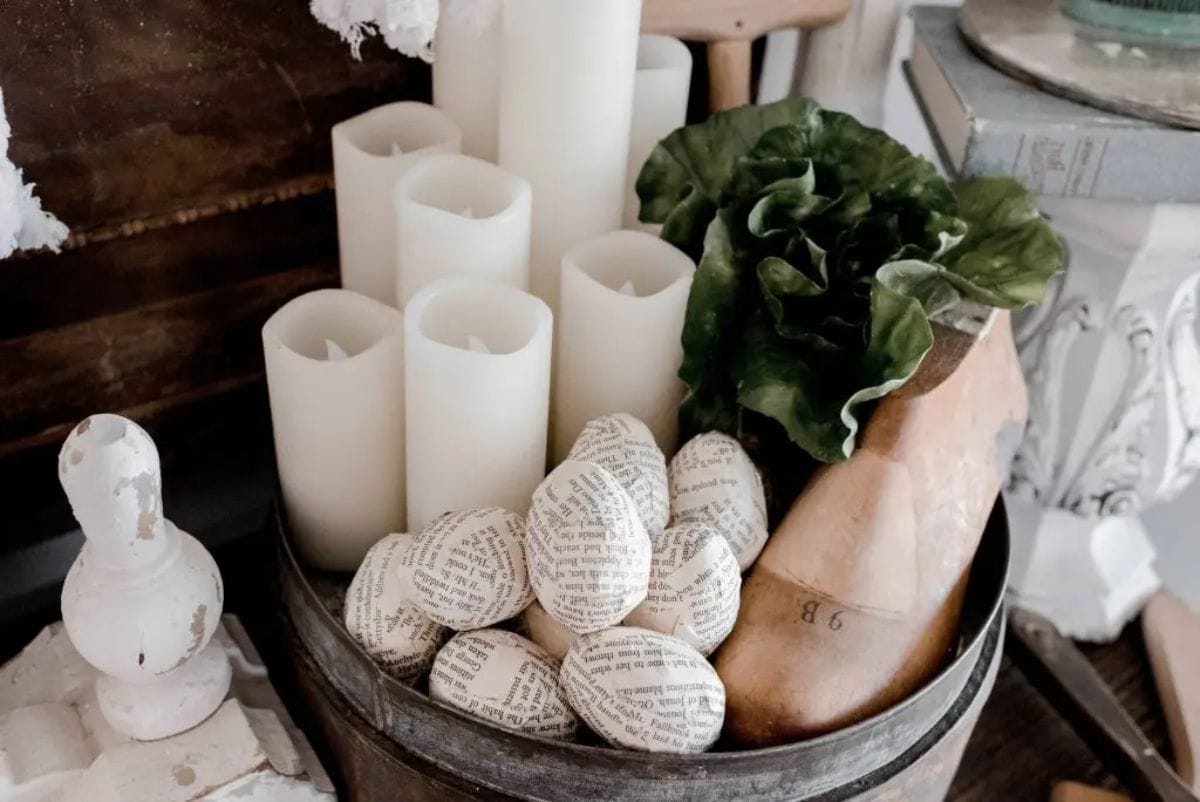 in a basket are white church cndles, green foliage and a pile of egg shapes covered in pages from old books