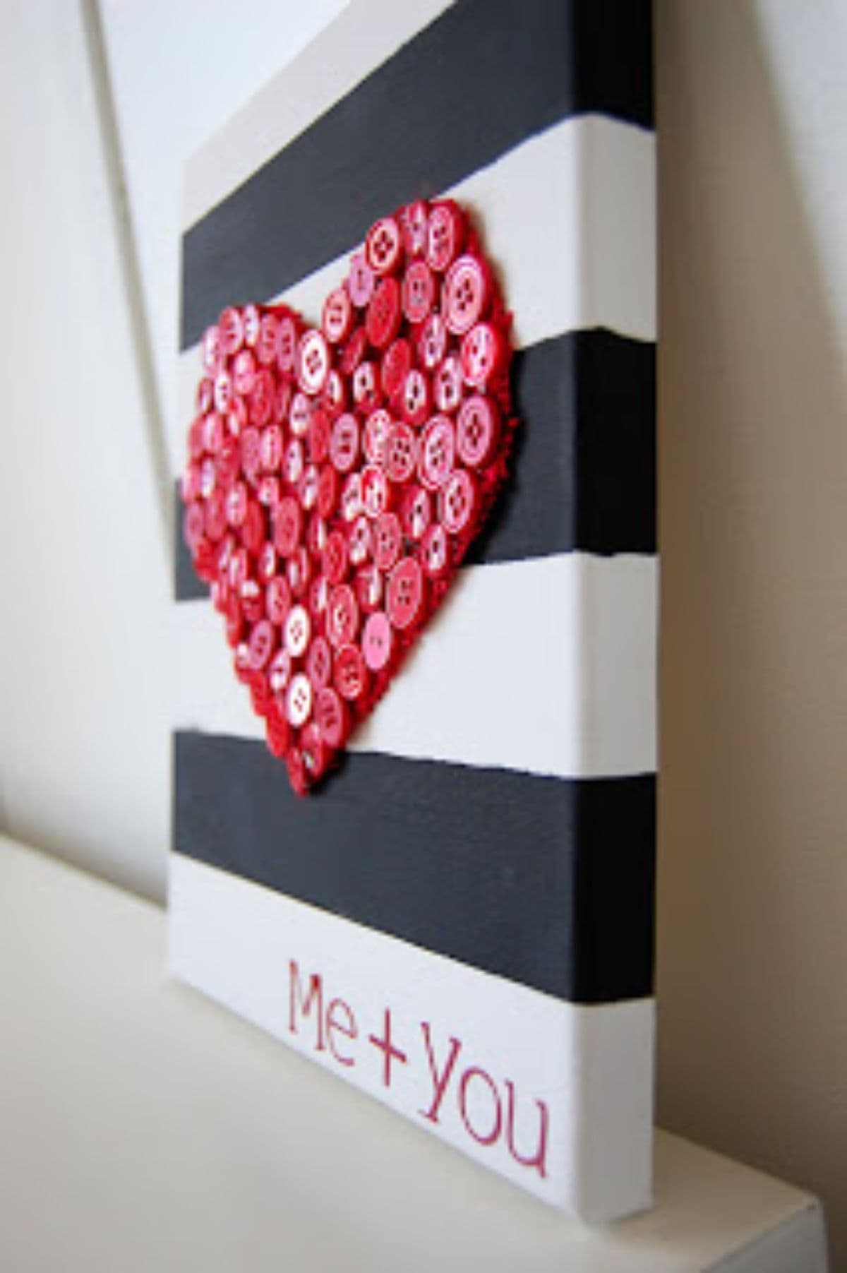 a side-on image of a blacj and white striped square hanging from a wall. On the front of the square is a heart shape made of red buttons. Underneath is the red text "ME + YOU"