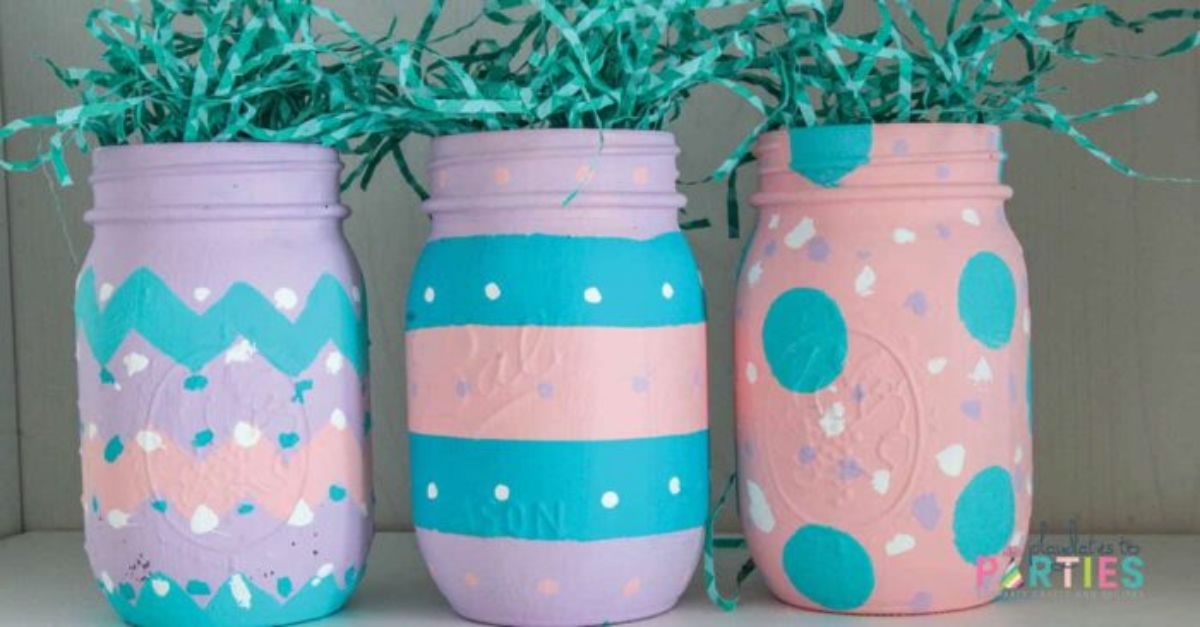 3 mason jars are decorated in pink, blue and white. The tops are stuffed with green shredded paper