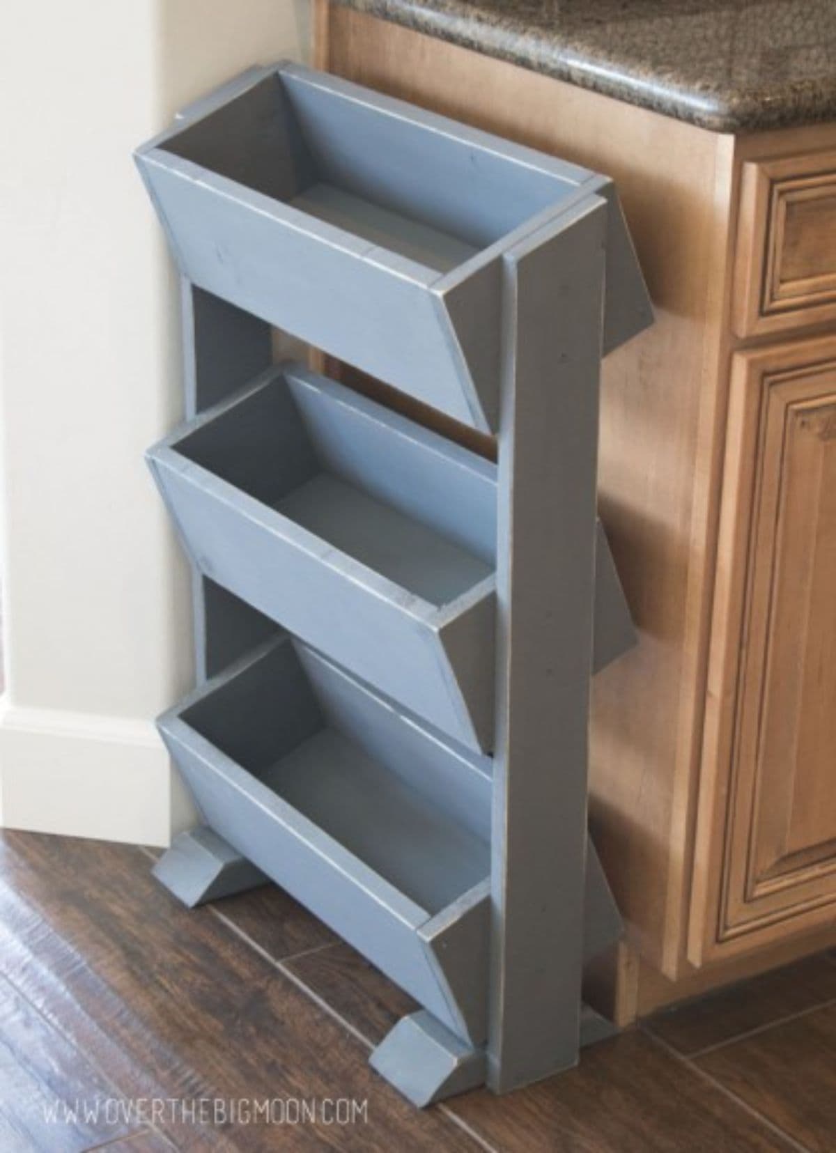 Sitting on the edge of a kitchen unit is a 3 tier vegetable display unti, with each grey wooden box shelf at a slant