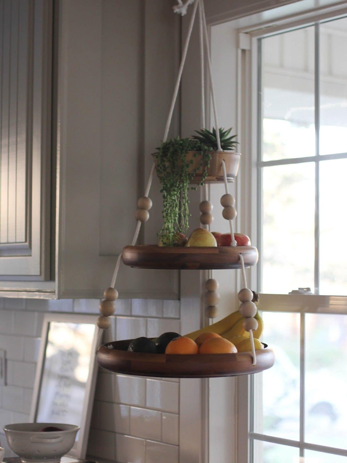 Hanging in front of a window is a 3 tier hanging shelf with fruit on each level