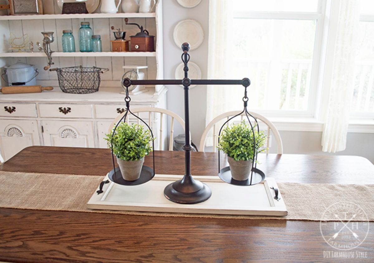 On a kitchen table is a white wooden tray. On top of it are some old weighing scales with a plant pot balanced on each side