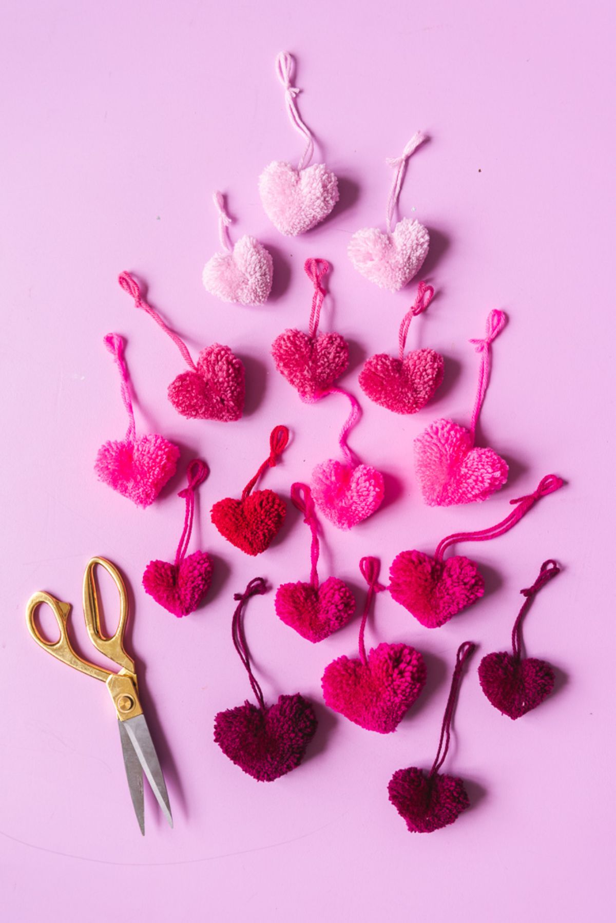 on a pink background are 16 knitted hearts graded from light pink to dark red. On the left is a pari of yellow handled scissors