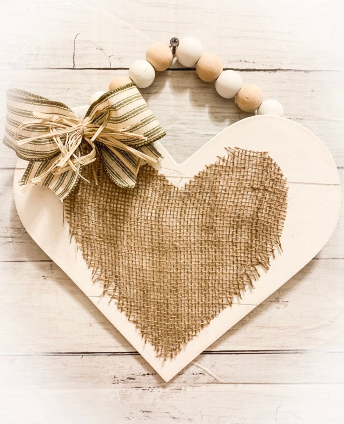 Hanging on a cream brick wall from a strong of wooden balls is a cream heart with a burlap heart stuck to the front.