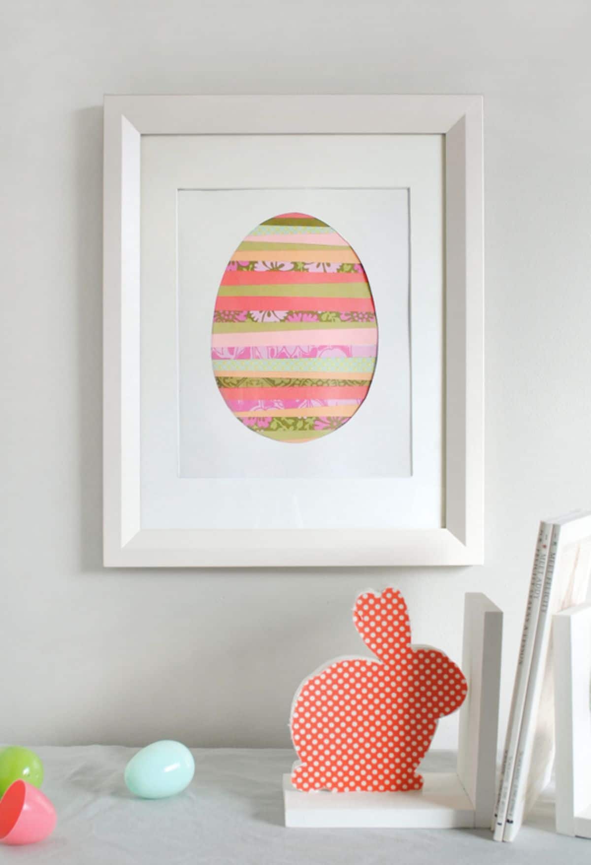 Hanging on a wall is a framed picture of a striped egg. In front sits a red bunny cutout