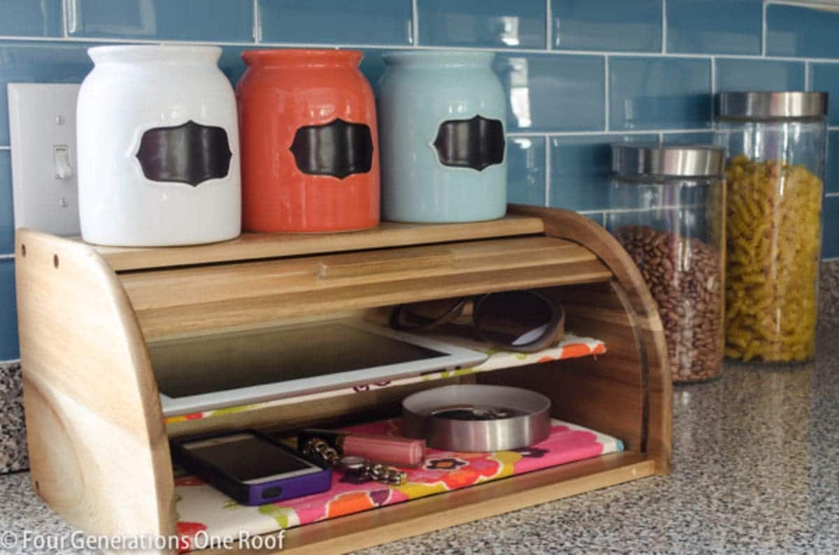 ON a countertop with blue subway tiles behind it is a wooden bread bin. On top sit 3 painted jars in white, orange and blue. A shelf has been put in the bread bin half way up and an ipad sits on teh top shelf with phones on the bottom