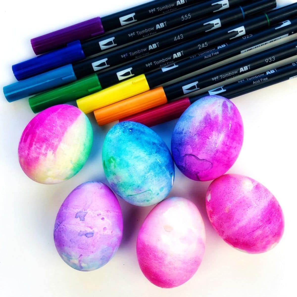 a pile of brush pens sit above six decorated hard boiled eggs on a white background