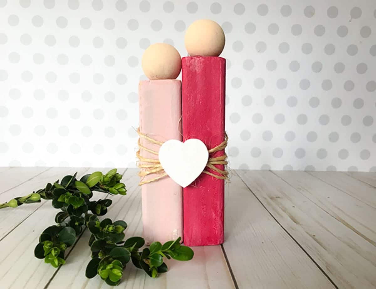 on a light wooden table, next to some green foliage is a scuplture of 2 people made of rectangular blocks of wood, one red, one pink. Wooden balls are their heads and they are tied together with twine and a white heart