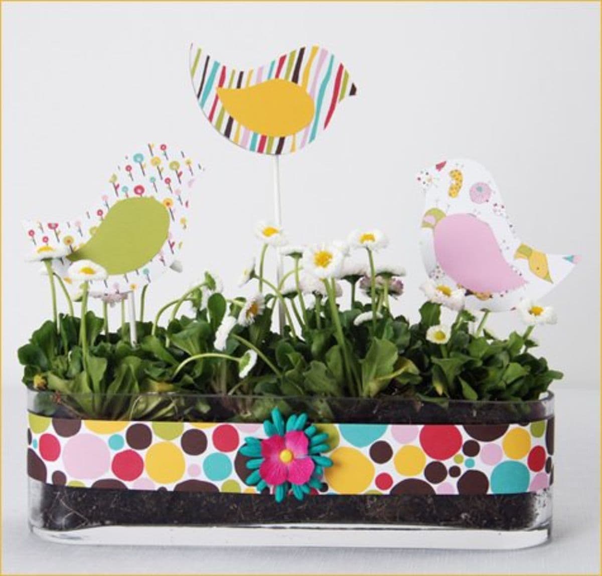 A clear tub is filled with green foliage. There is a spotty ribbon wrapped aroun dit and sticking out of the top are 3 paper birds