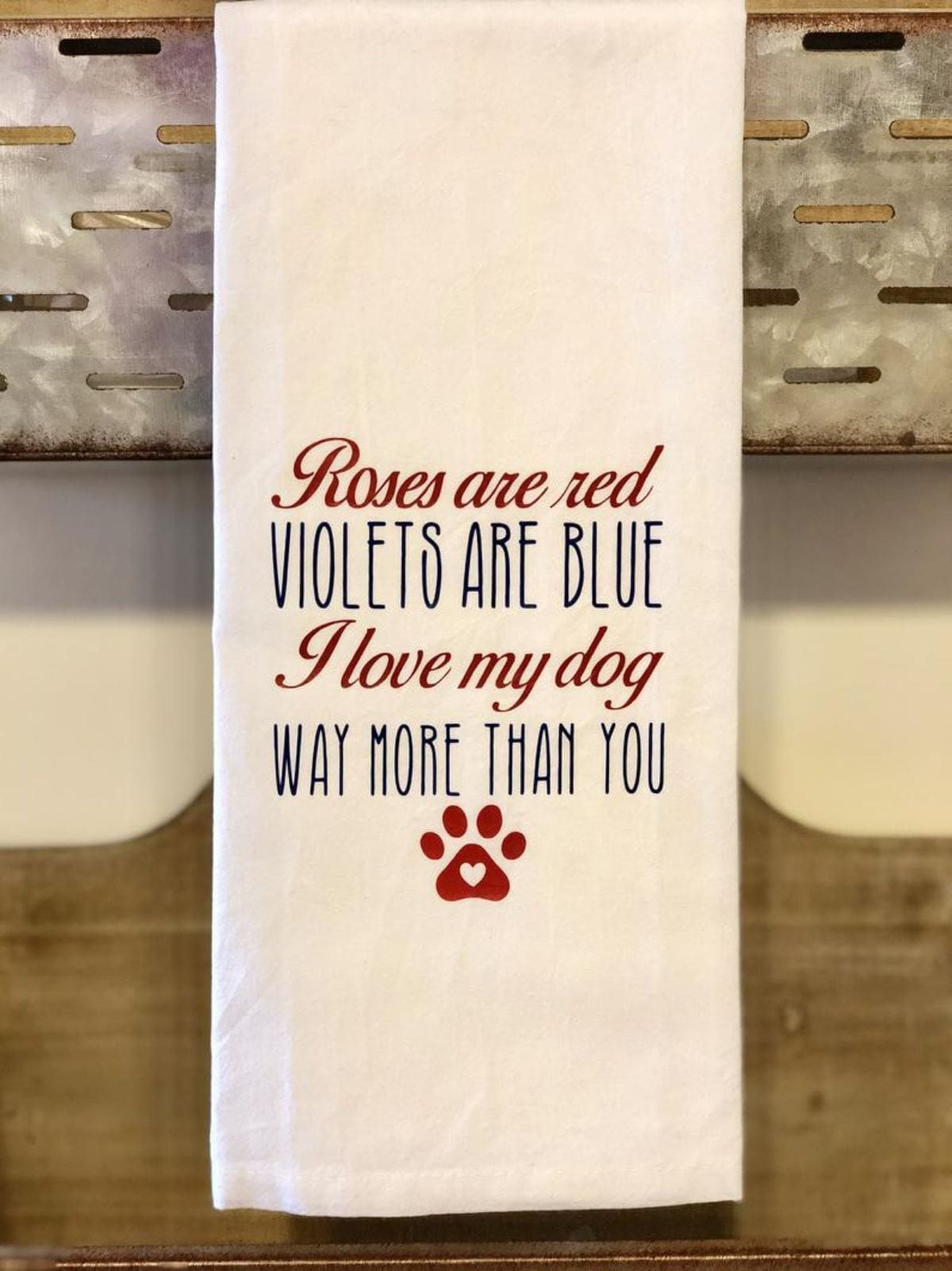 Resting on the top of a wooden cabinet is a cream tea towel with the red and black text "Roses are red, Violets are blue, I love my dog, whay more than you". UNderneath is a red paw print