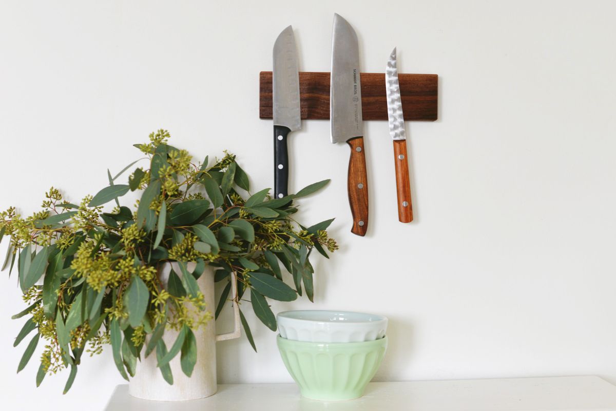 On a white wall is a wooden rectangle. Stuck to it are 3 different sized knives. In front is a potted plant in a white pot and a small bowl.