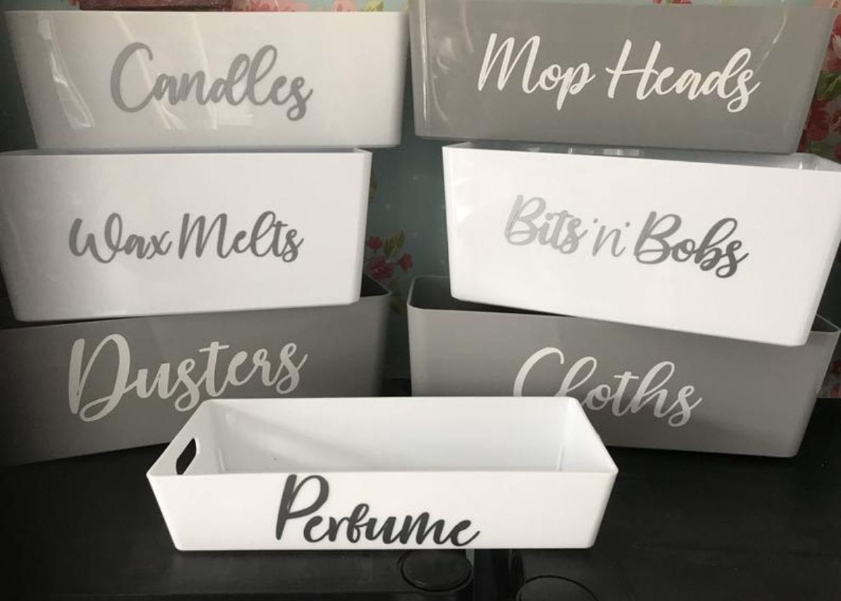 2 stacks of grey and white plastic tubs with swirly lettering on them: candles, wax melts, dusters, mop heads, bits n bobs, cloths, perfume