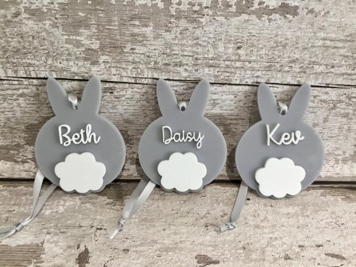 on a background of wooden planks, sit 3 grey bunnies with white tails and names on each of their backs