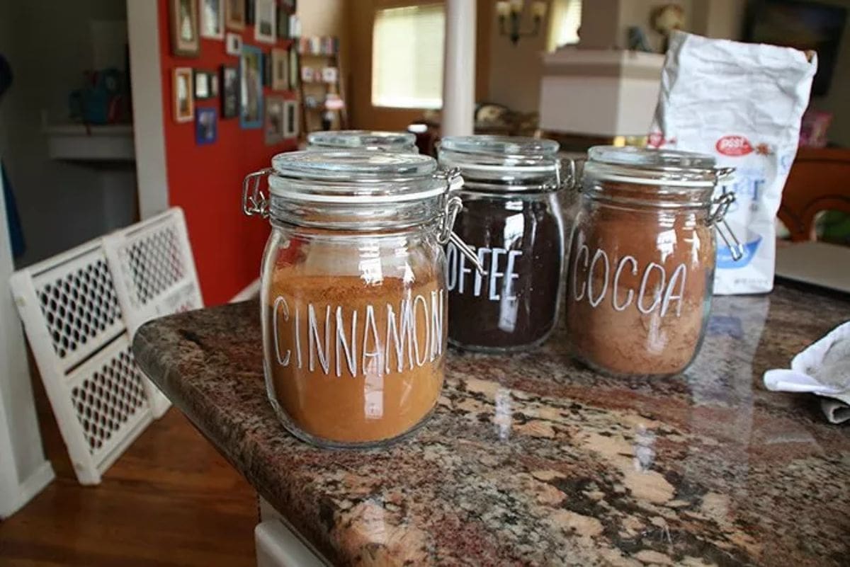 On a marble countertop sit 3 glass mason jars. They have chalk writing on them labelling their contents: Cinnamon, Coffee adn Cocoa.