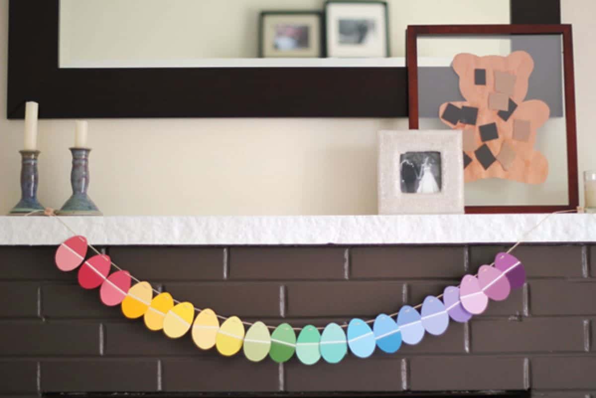 a garland of colored egg shapes hang on a mantelpiece decoratied with a mirror and pictures