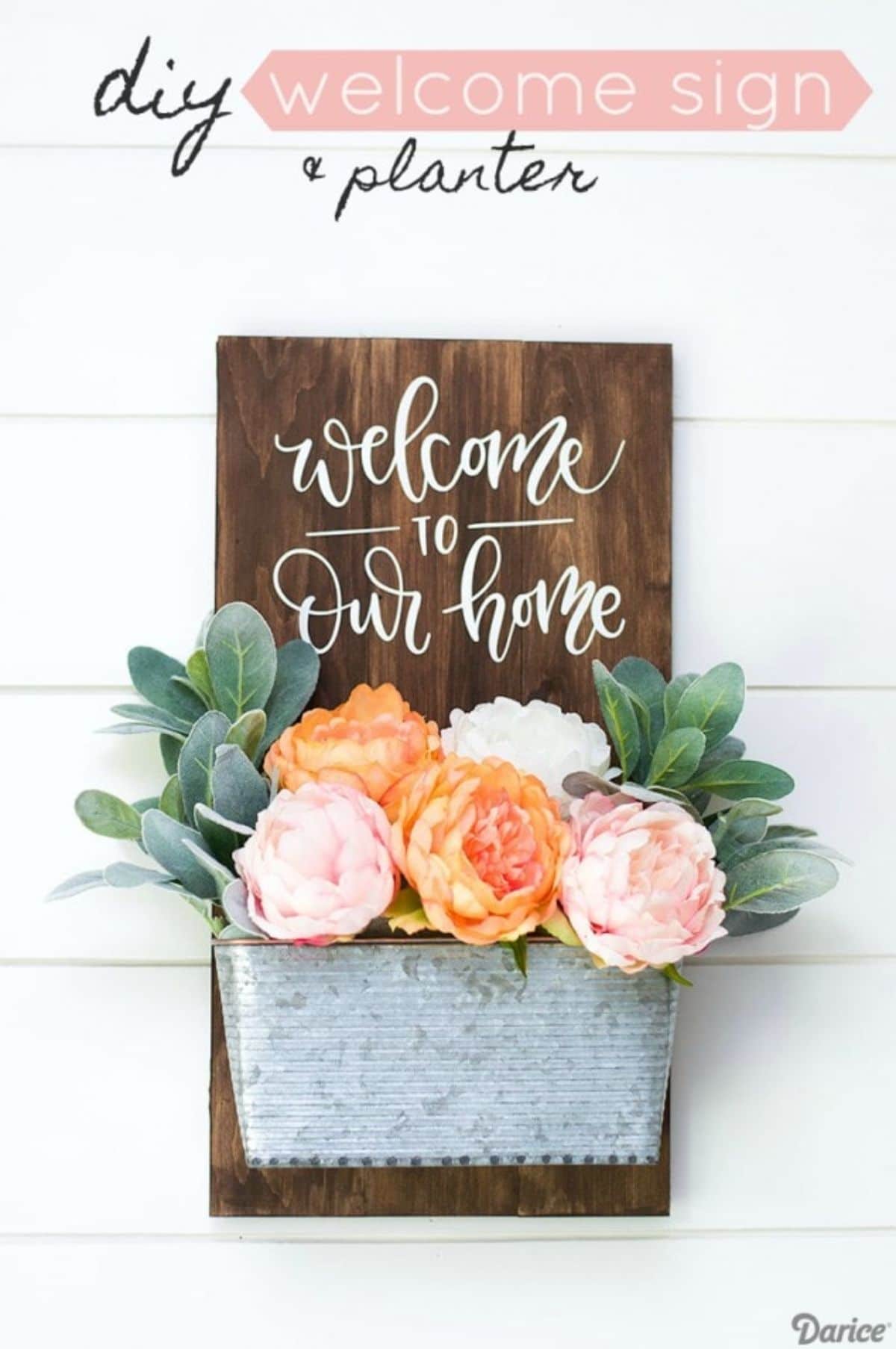 On a white background is a dark wooden board with "Welcome to our house". Stuck to the front of it is a metal tub filled with orange and pink flowers