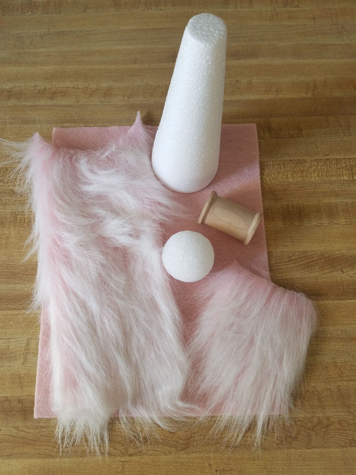 On a wooden table sits a pink ssquare of fabric. On top of the fabric are 2 pieces of faux fur, a ping pong ball, a reel of cotton and a yarn cone