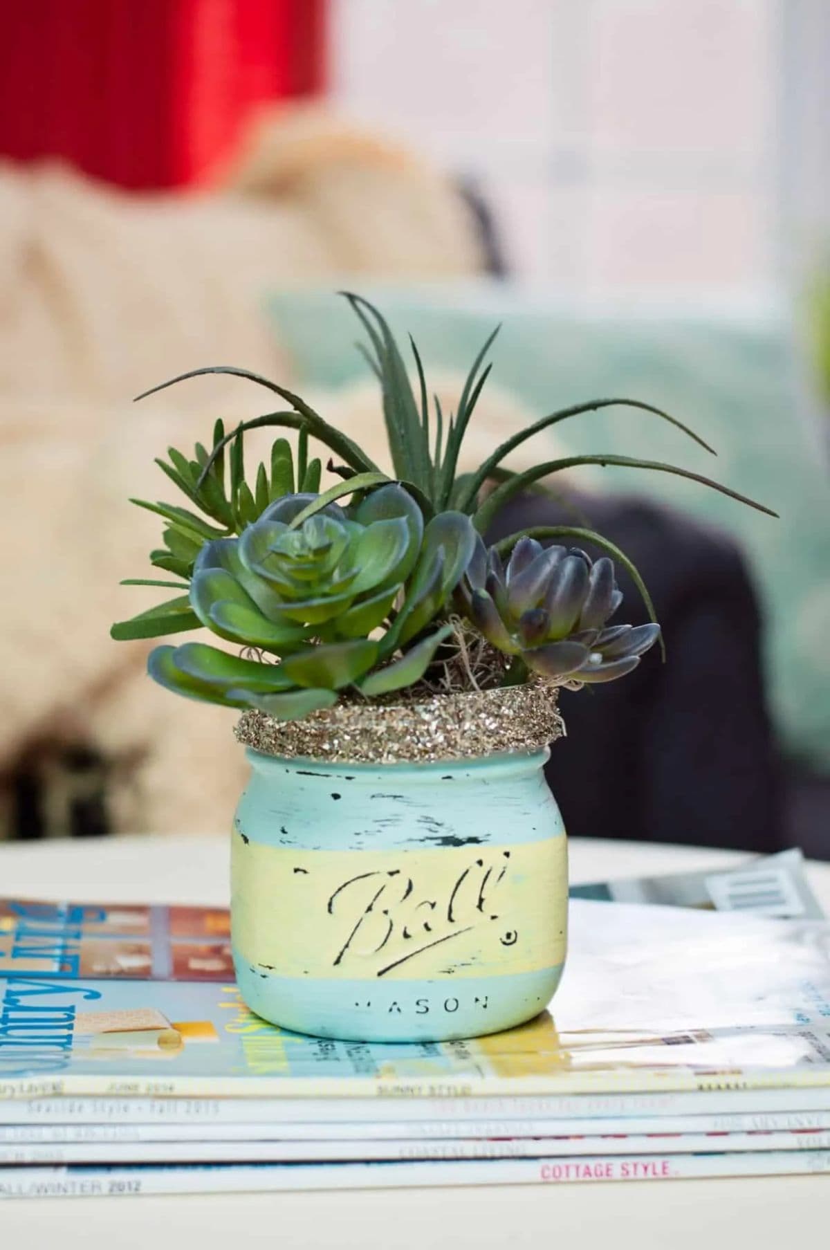 A mason jar painted in blue and green stripes sits on a table. It is filled with a succulent plant