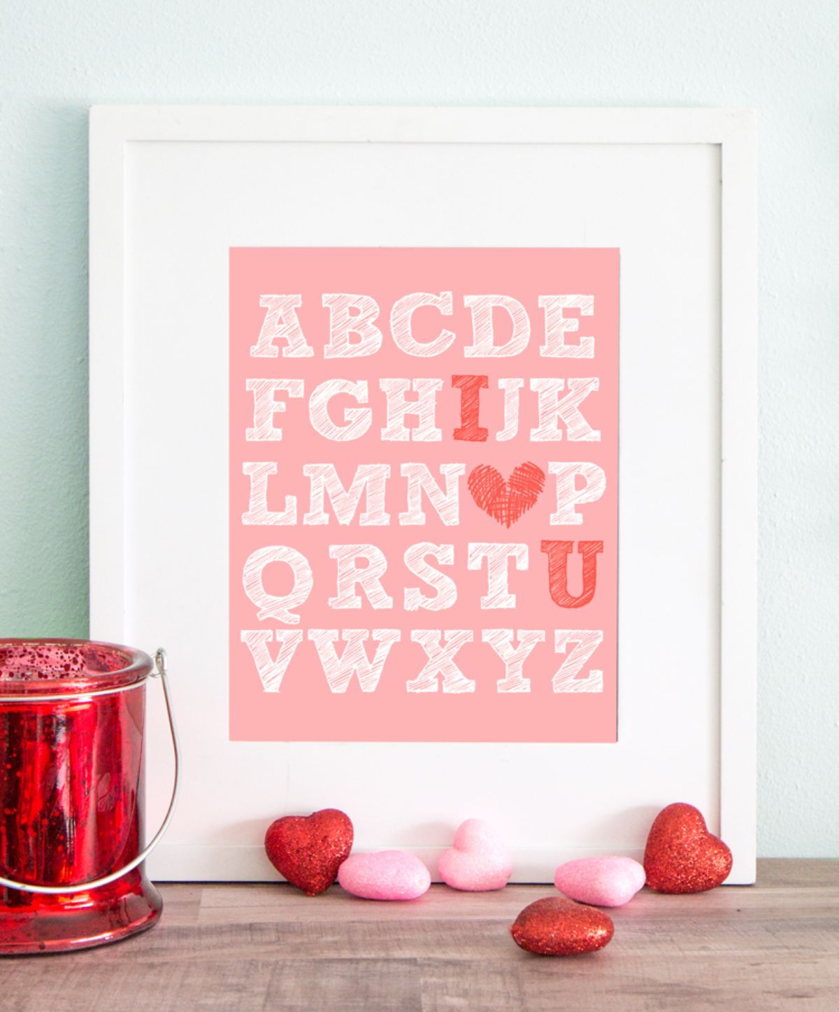 A white framed print sits on a wooden table. The background of the print is pink and the alphabet runs down the page. I and U are in red, and O is shaped like a red heart.